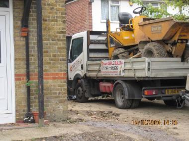 digger on flatbed driving down the side of house