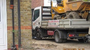 digger on flatbed driving down the side of house